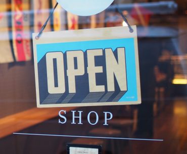 An open sign hangs from a glass door to a business directly above the word “shop.”