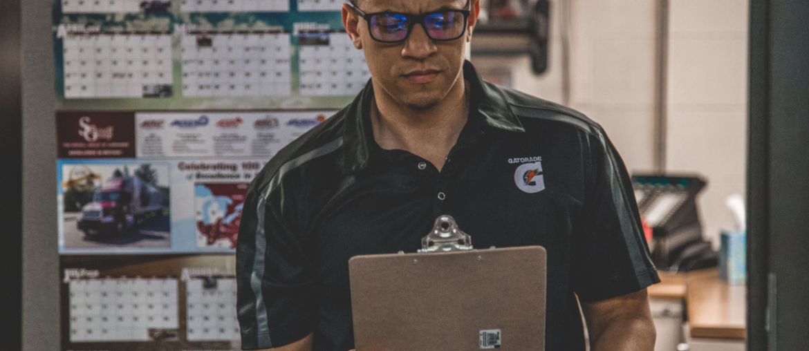 Man holding a clipboard with a serious look on his face and multiple calendars hanging on the wall in the background.