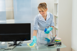 A cleaning employee wearing a traditional housekeeper dress is using a cleaning solution and cloth to wipe down an office desk in a brightly-lit room.