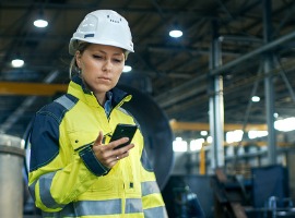 female industrial worker in the hard hat uses mobile phone while walking through heavy