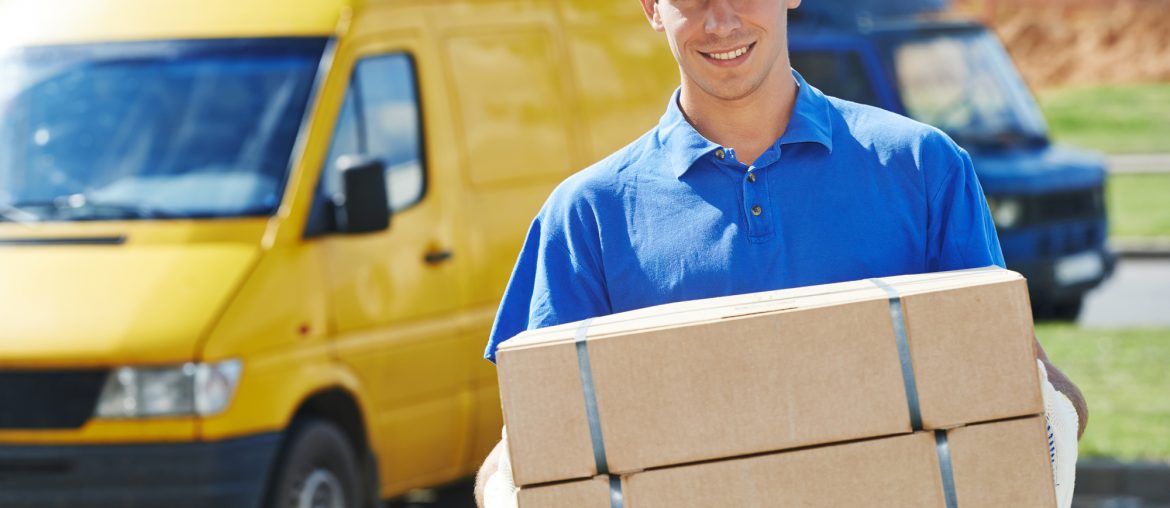 A delivery driver dressed in a blue polo shirt and a blue hat is holding two cardboard boxes. They are standing in front of a yellow delivery van.