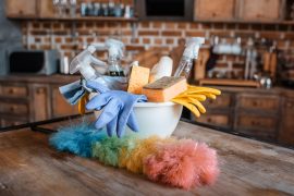 A white bowl full of cleaning supplies sits on top of a dusty wooden table. In the foreground, there is a rainbow colored duster. In the background, a wooden kitchen counter with various appliances is visible.