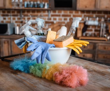 A white bowl full of cleaning supplies sits on top of a dusty wooden table. In the foreground, there is a rainbow colored duster. In the background, a wooden kitchen counter with various appliances is visible.