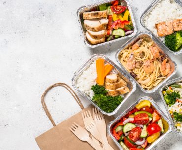 Overhead view of several individual meals packaged in tin foil containers, next to a brown paper delivery bag and several plastic forks.