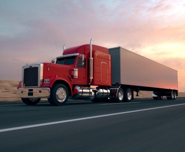 A red semi-truck with a white trailer is driving on the highway. Dry terrain and a slightly cloudy sunset are visible in the background.