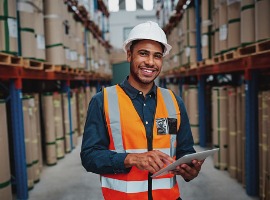 happy male factory manager using digital tablet in warehouse while standing against goods