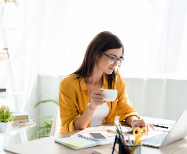 A person sits at their desk, sipping coffee as they work on their laptop from home.