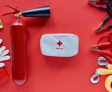 Overhead view of a white first aid kit surrounded by tow cable, jumper cables, gloves, a vest, and a fire extinguisher.