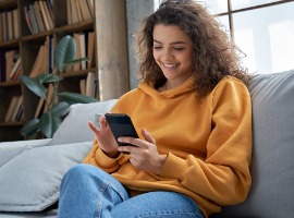 happy millennial hispanic teen girl checking social media holding smartphone at home smiling